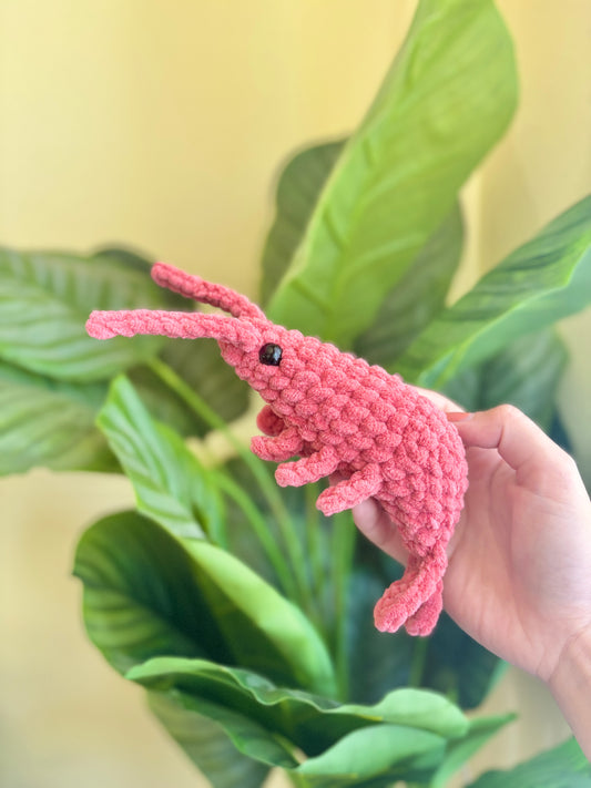 Hand holding a red crochet shrimp toy with green leaves in the background