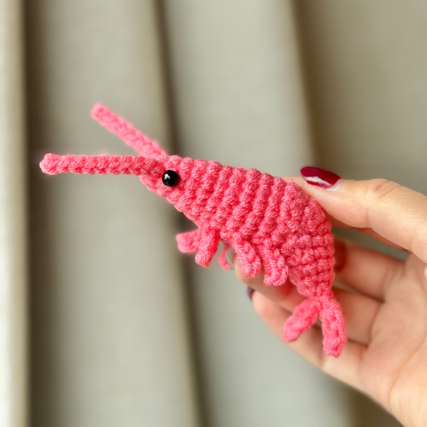 A hand holding a pink crochet shrimp against a neutral background