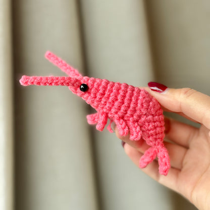 A hand holding a pink crochet shrimp against a neutral background