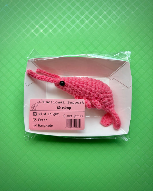 Pink crocheted shrimp plushie with an emotiona support shrimp label on a green background