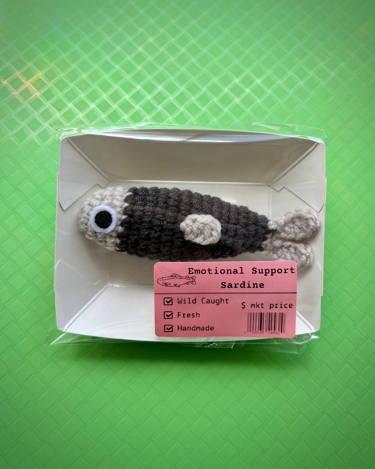 Crocheted sardine plush in packaging with a emotional support sardine label on a green textured background