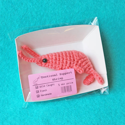 Pink crocheted shrimp with an "emotional support shrimp" label on a turquoise background