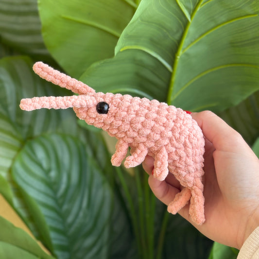 Hand holding a pink plush crochet shrimp against a green background with leaves
