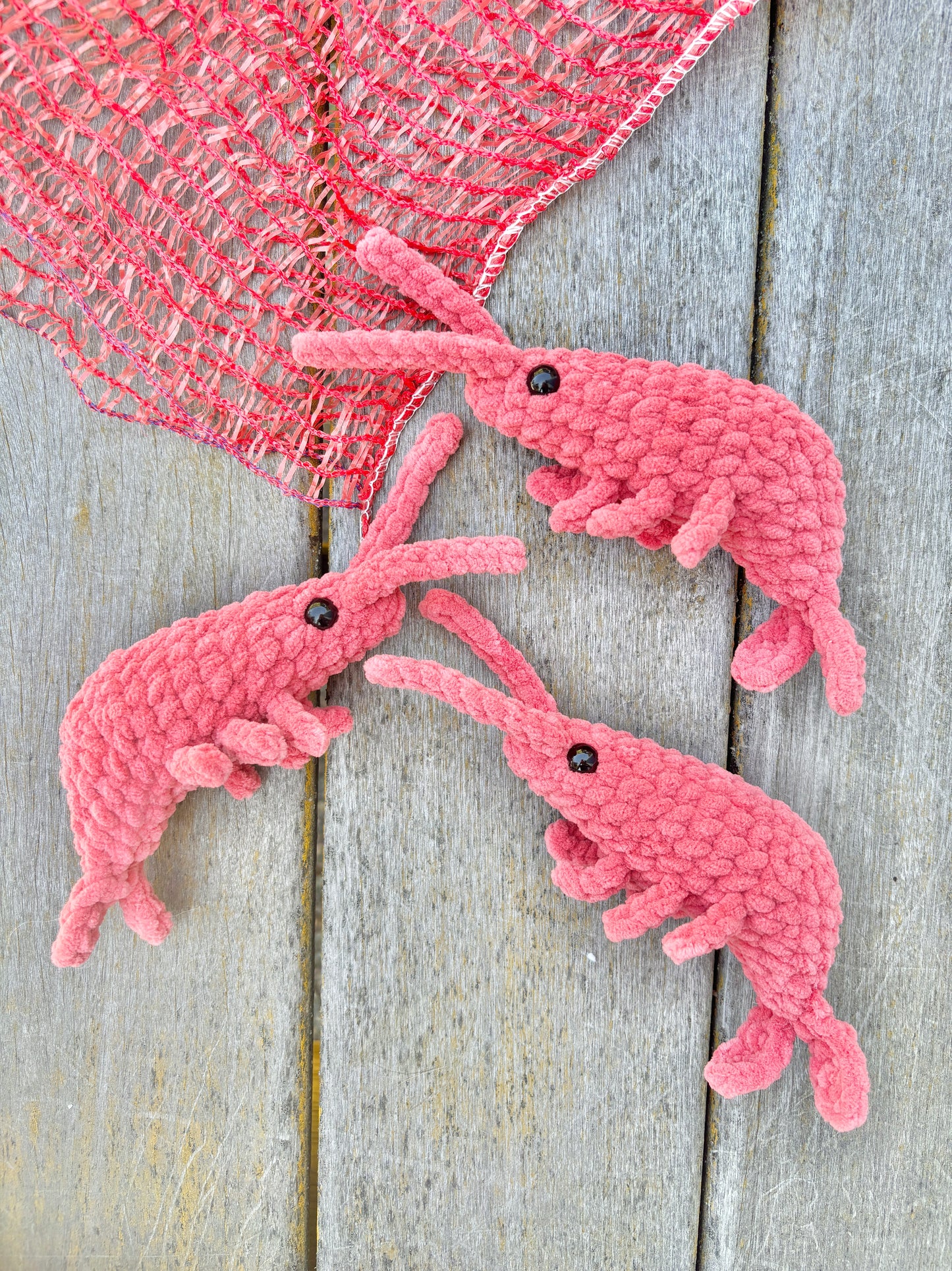 Three pink crochet shrimp on a wooden surface with a pink fishing net.