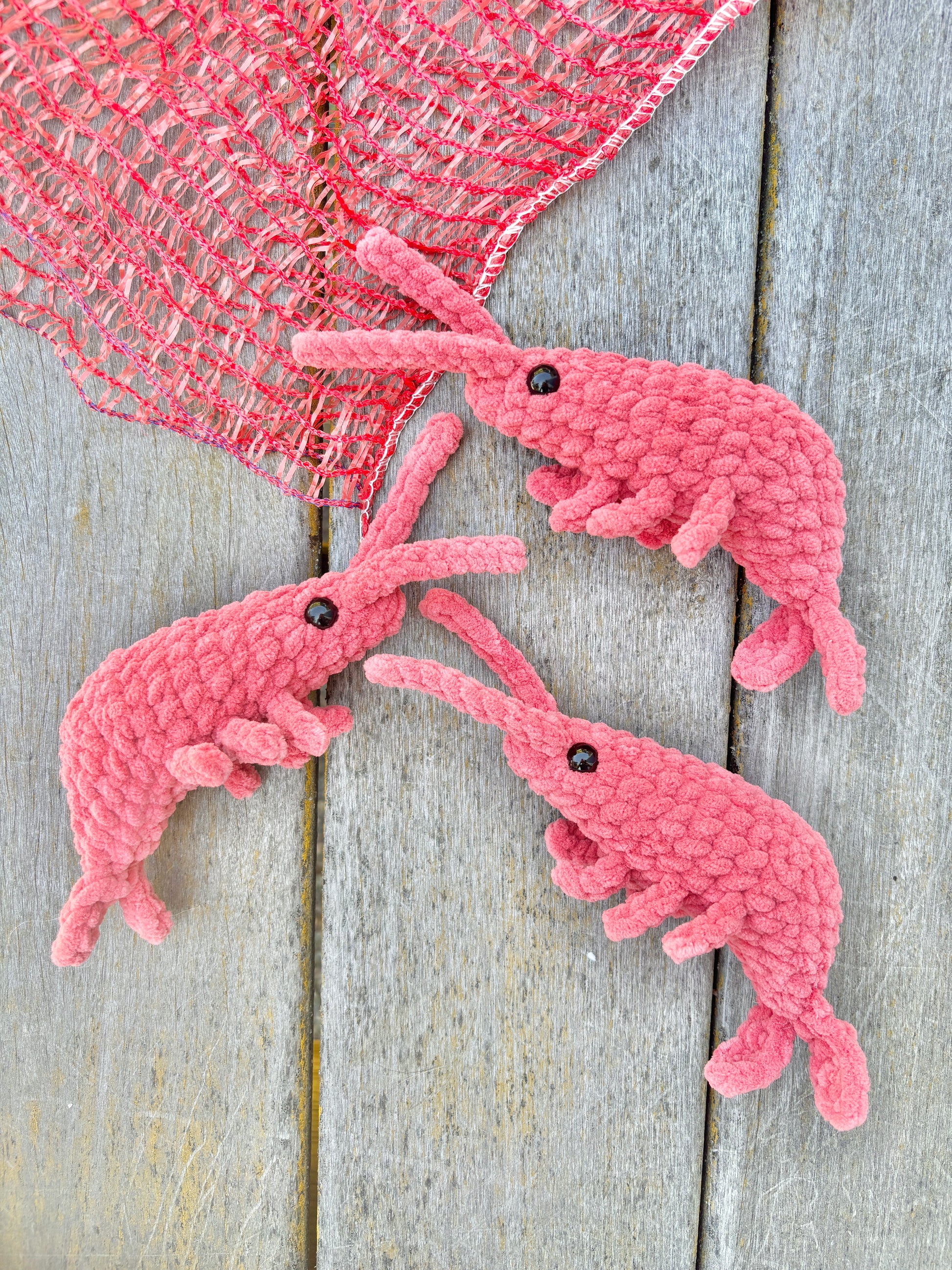 Three pink crochet shrimp on a wooden surface with a pink fishing net.