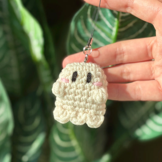 Hand holding a small crocheted ghost keychain with a leafy background