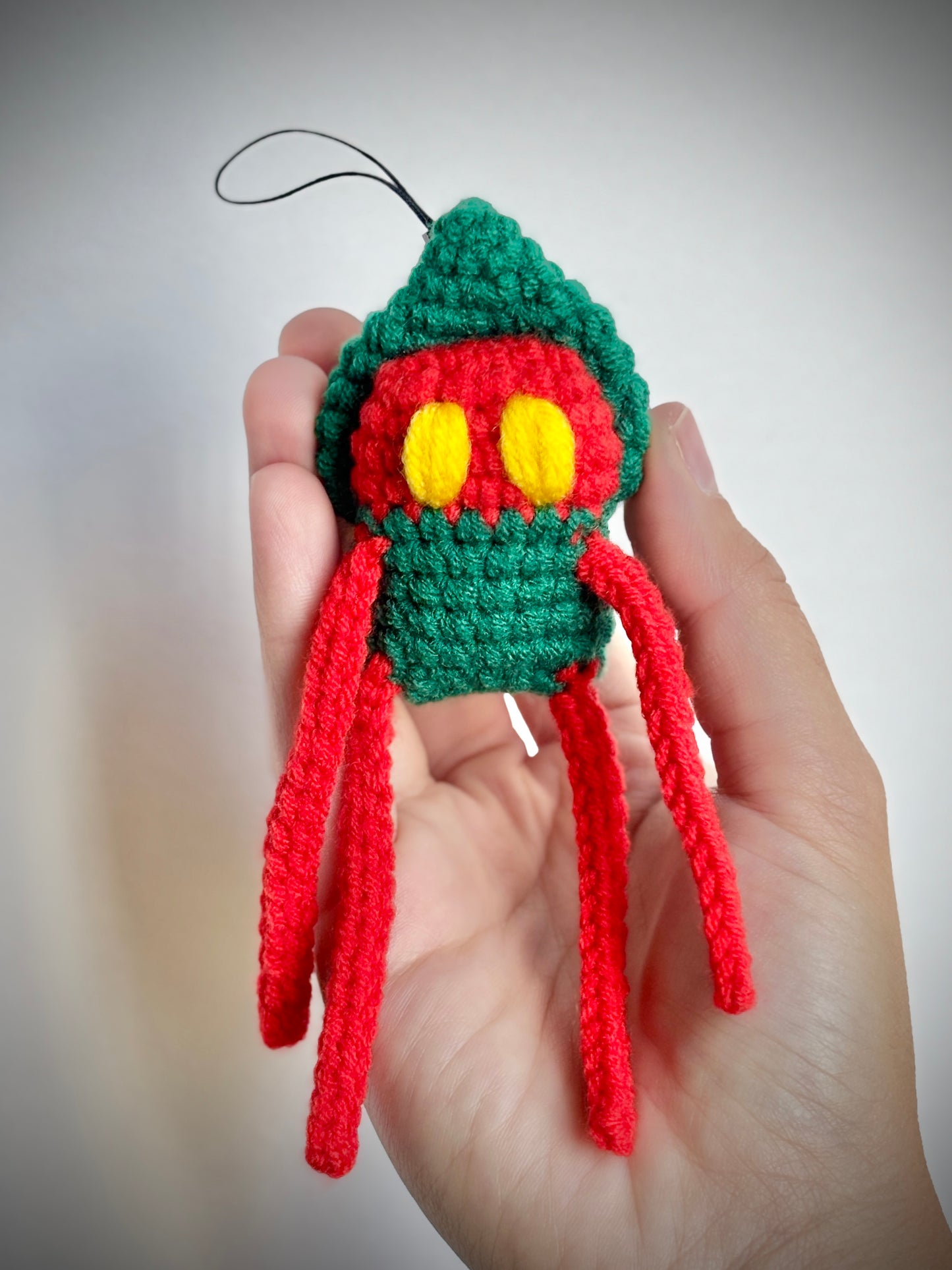 Hand holding a small crocheted green and red creature with yellow eyes against a neutral background