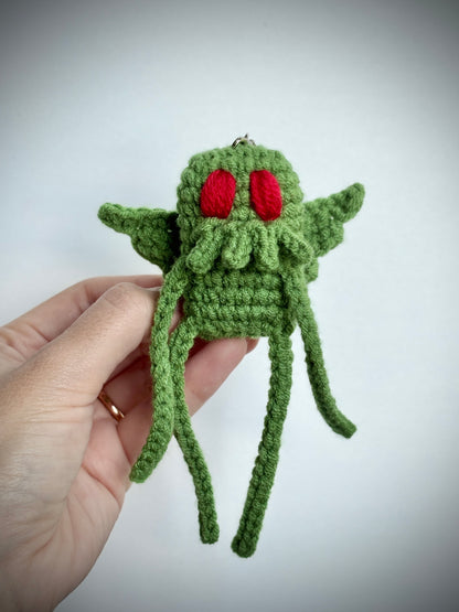 Green crocheted creature with red eyes held by a hand against a light gray background