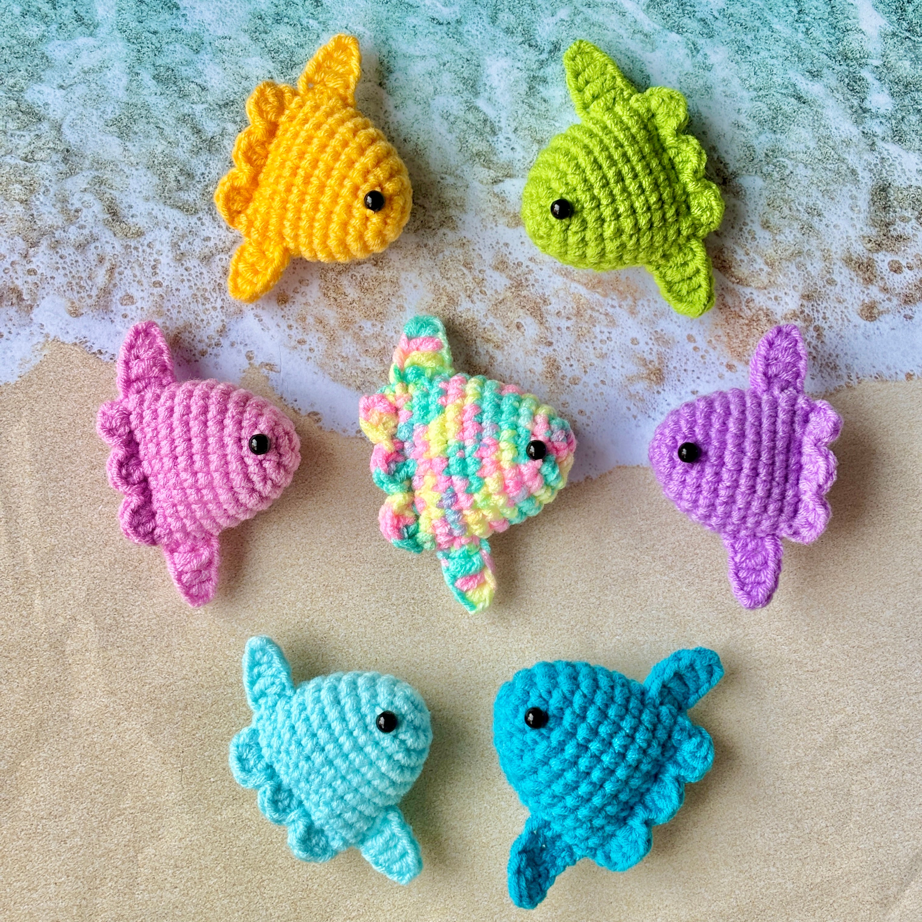 Colorful crocheted fish toys on a sandy surface with water in the background