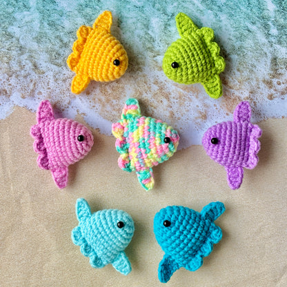 Colorful crocheted sunfish plushies on a sandy surface with water in the background