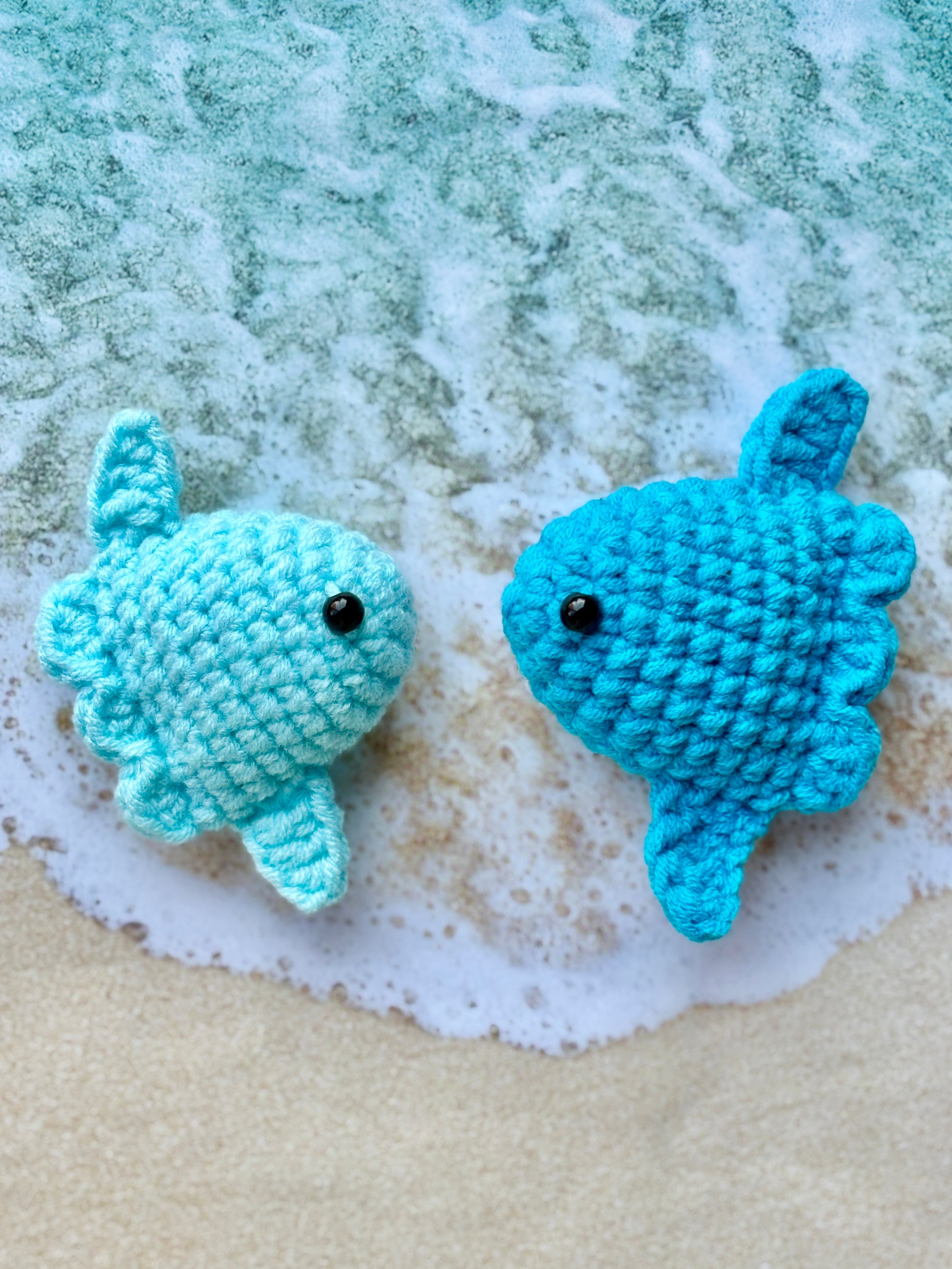 Two blue crochet fish on a sandy surface with water.